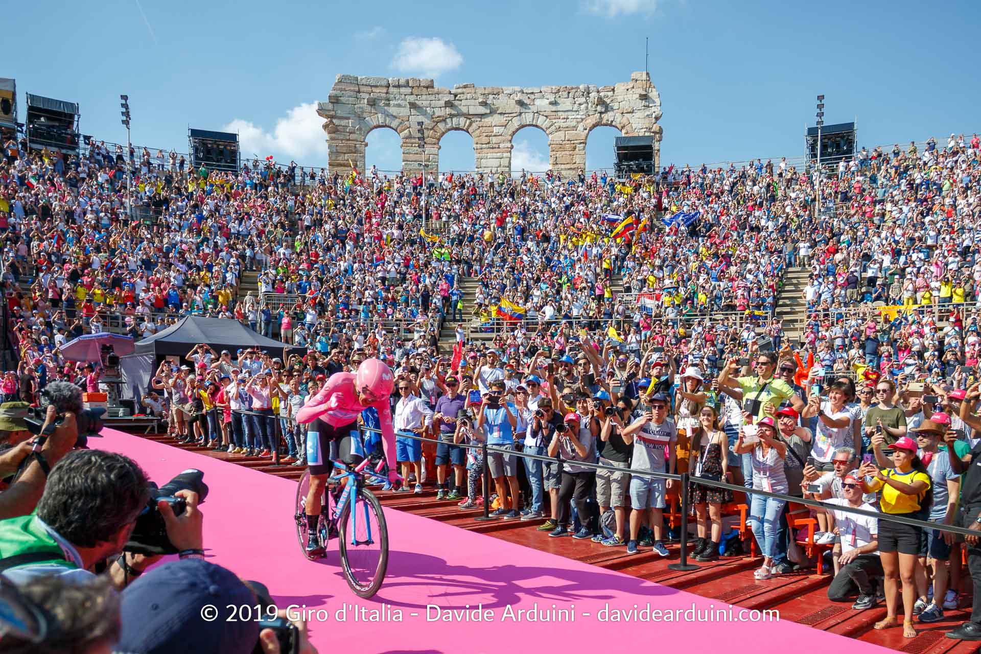 Ciclista in maglia rosa al Giro d'Italia, circondato da una folla in un'arena con bandiere e striscioni.